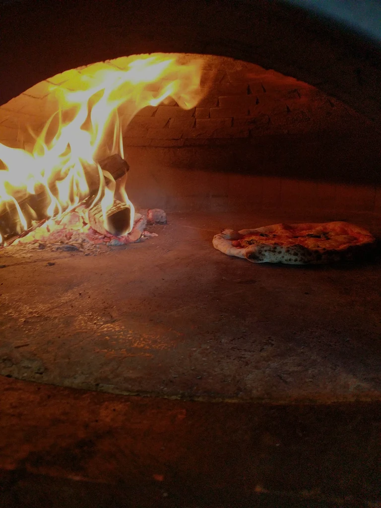 Pizza baking in a brick wood-fired oven next to a fire.