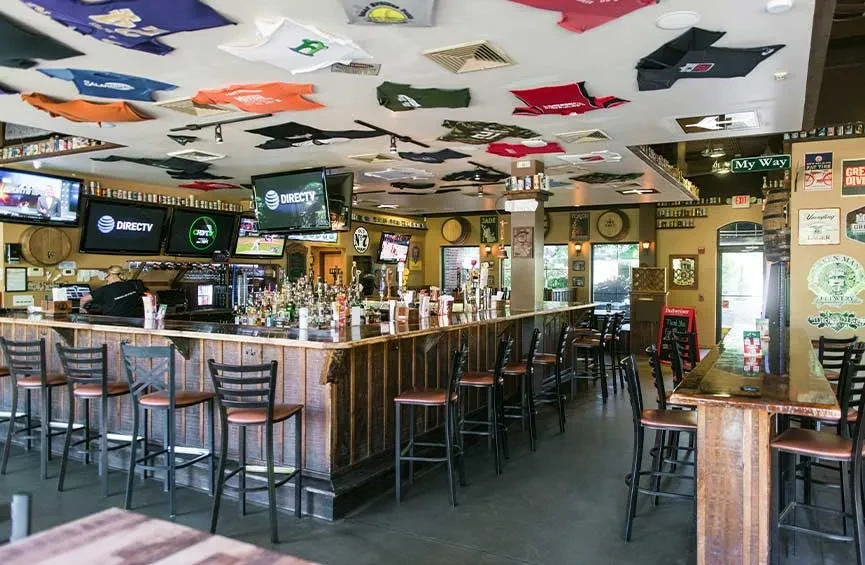 L-shaped bar in a sports pub with TVs, liquor, and t-shirt ceiling.