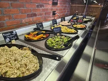 Stainless steel buffet line with labeled salads and roasted vegetables.