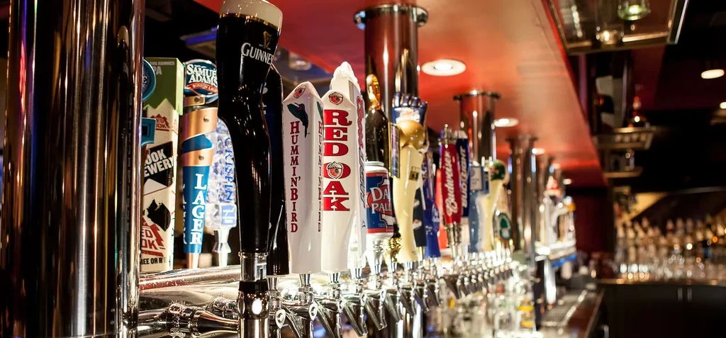 Long row of metallic beer taps with branded handles in a bar.