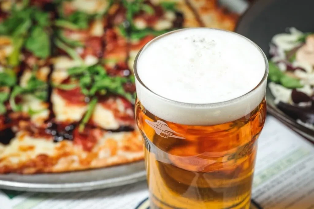 Close-up of beer pint with foam, blurred pizza and salad.
