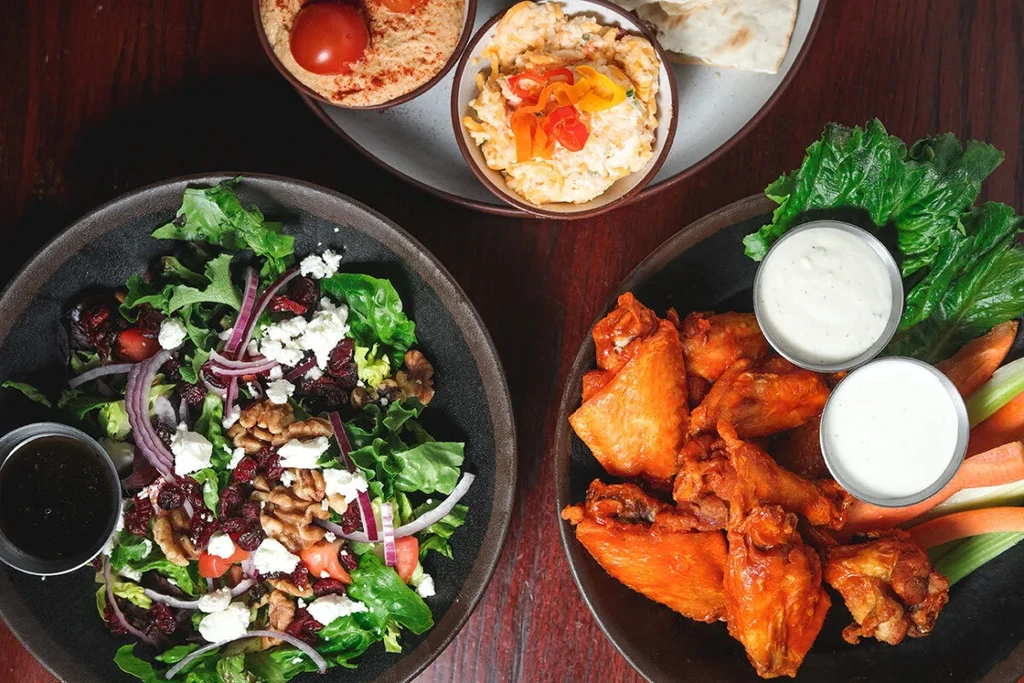 Close-up overhead of colorful salad, chicken wings, dips, and flatbread.