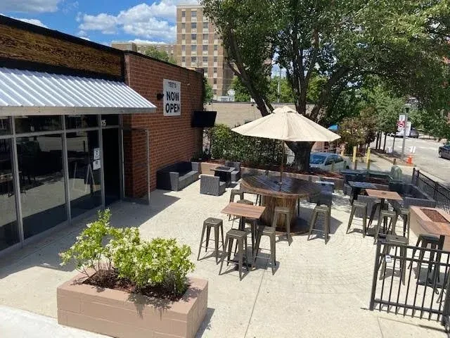 Brick building with 'TEES NOW OPEN' sign and sunny outdoor patio.