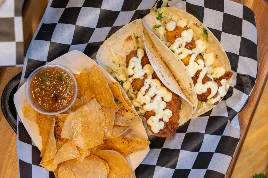 Two crispy fish tacos, chips, and salsa on a checkered liner.