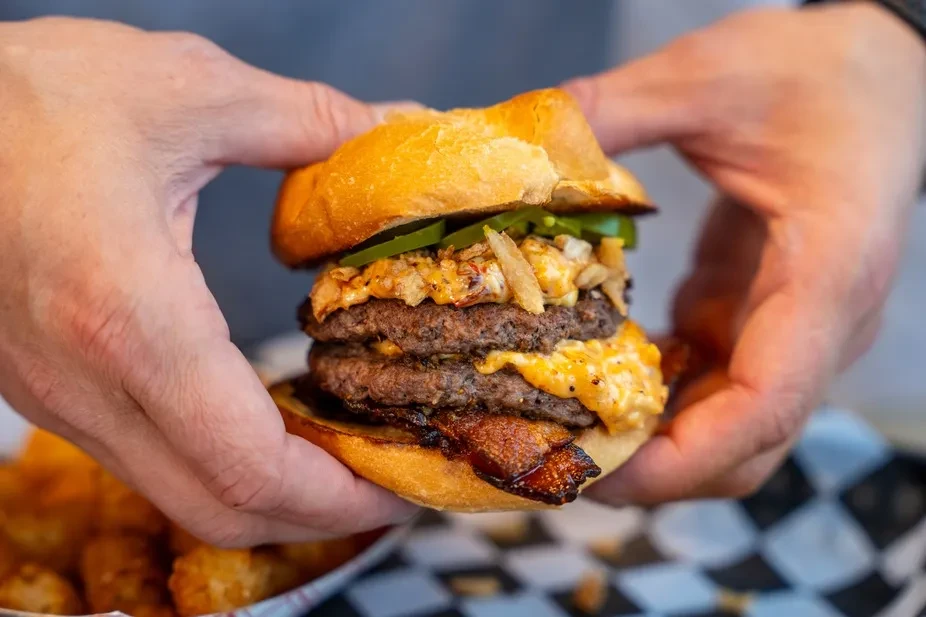 Hands holding a multi-patty burger with cheese, bacon, jalapeños, crispy onions.