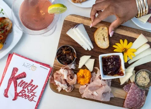 Overhead shot: Hand reaches for bread, charcuterie, cocktail, Kinky Boots program.
