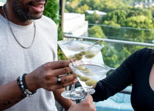 Two people toast three olive-garnished martinis outdoors.