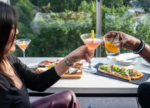 Two people toast cocktails and charcuterie at an outdoor restaurant.