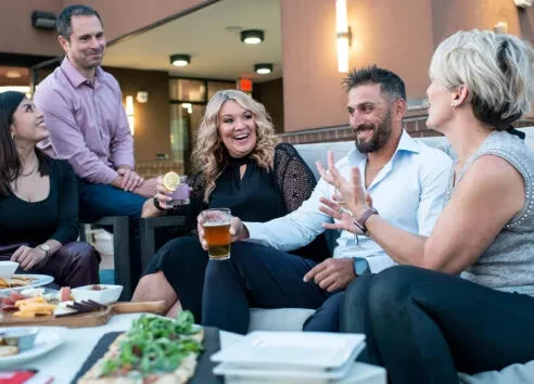 Five adults smiling on a restaurant patio with food and drinks.