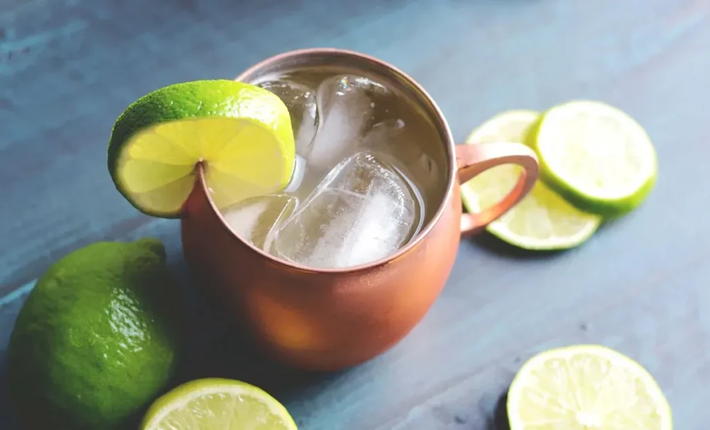 Copper mug drink with ice and lime wedges on a blue surface.