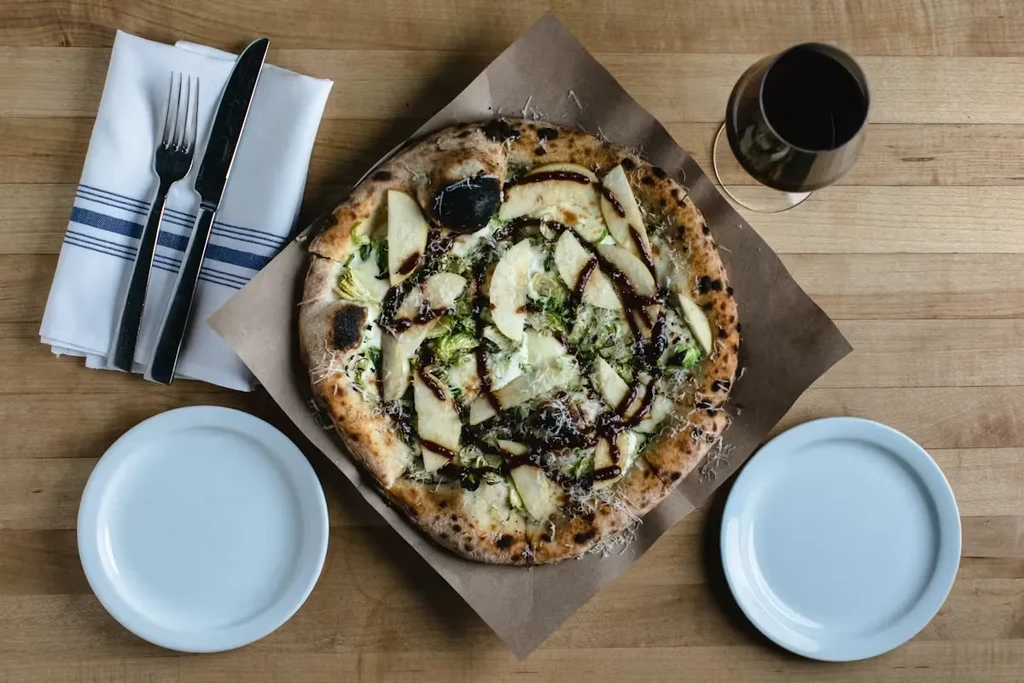 Gourmet pizza with fruit, greens, cheese, dark sauce, wine on wooden table.