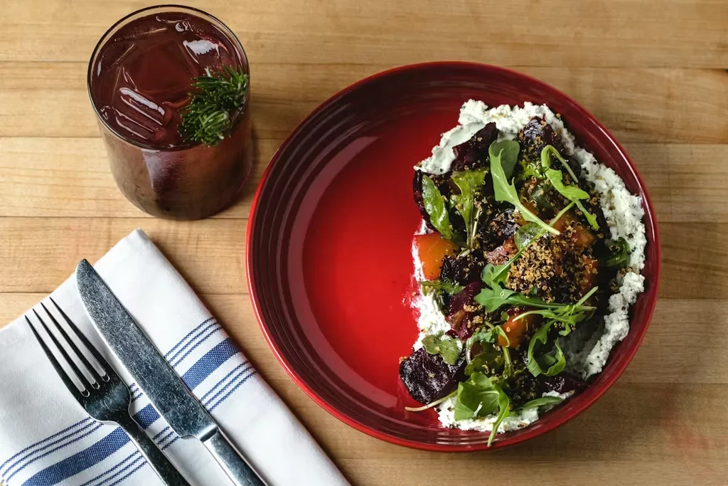 Plated beet and cheese salad with greens, drink, and cutlery.