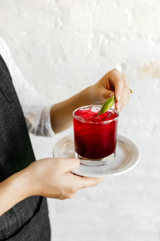 Hands garnish a red drink with ice and a lime wedge.