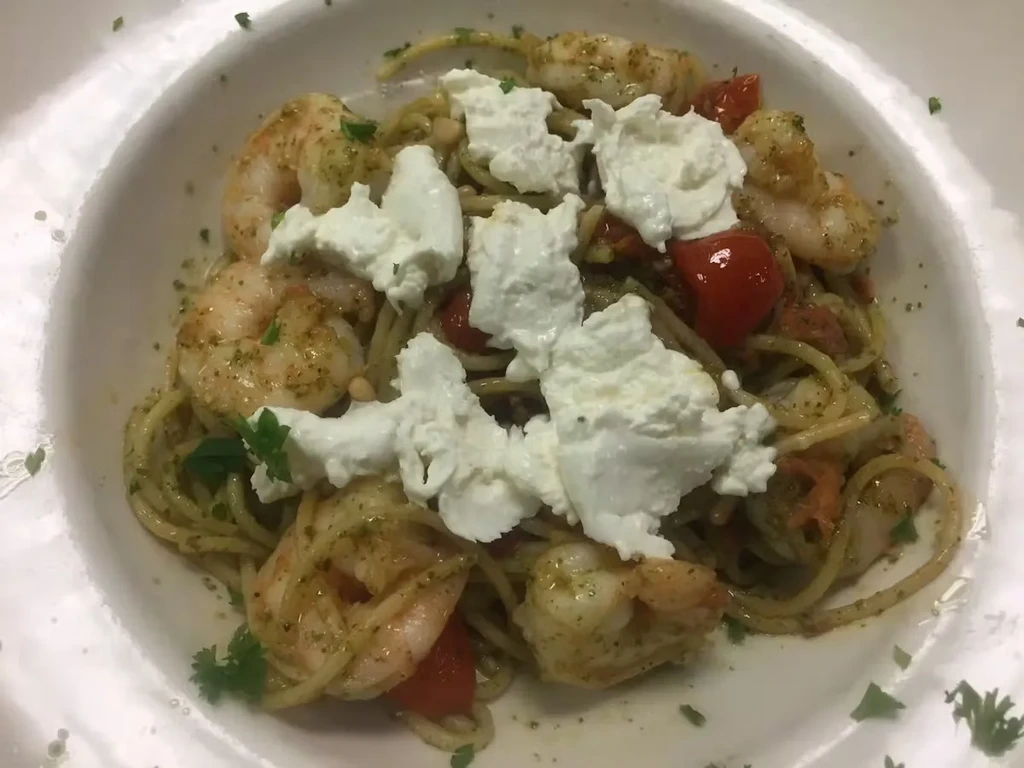 Pesto spaghetti with shrimp, tomatoes, cheese, and herbs in a white bowl.