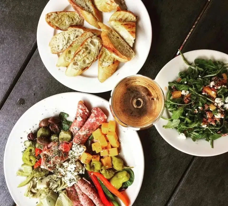 Overhead: Plates of charcuterie, salad, bread, and wine on dark table.