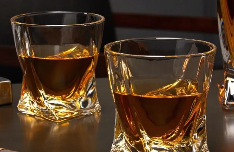 Two modern twisted glasses with amber spirits on a dark table.