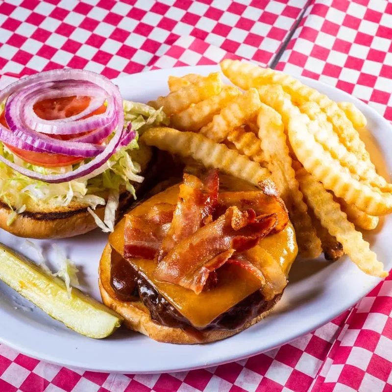Gourmet open-face bacon cheeseburger, fries, and pickle on checkered tablecloth.