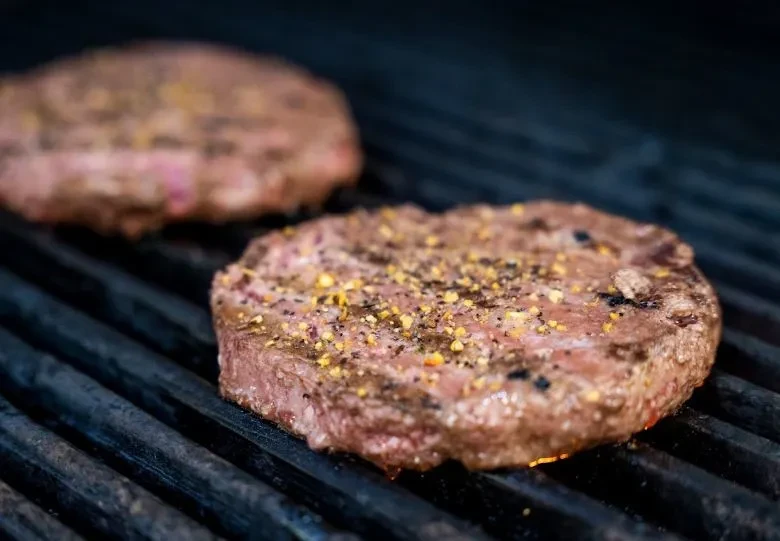 Two seasoned hamburger patties grilling on a dark grate with grill marks.