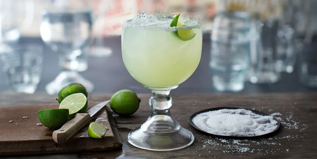 Salted margarita with lime, crushed ice, surrounded by limes and salt on wood.