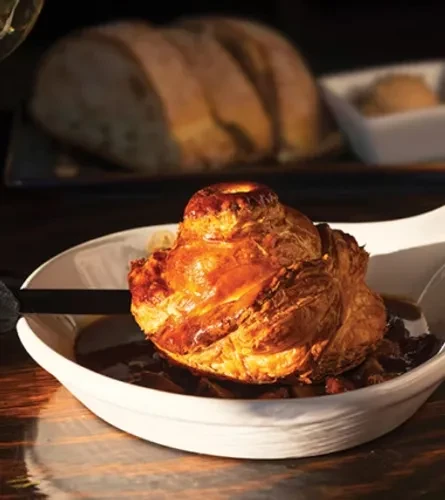 Golden-brown flaky pastry in dark stew, white boat dish.