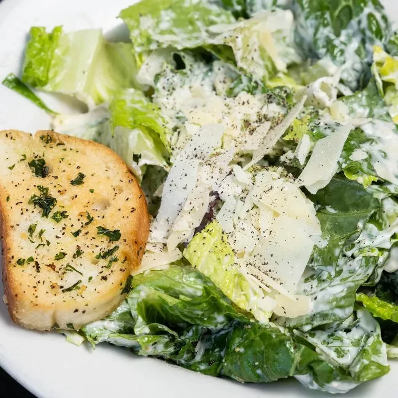 Close-up of Caesar salad with creamy dressing, Parmesan, and garlic bread slice.