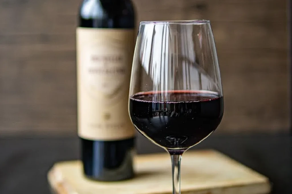 Close-up of a red wine glass with bubbles, blurred bottle on wood.