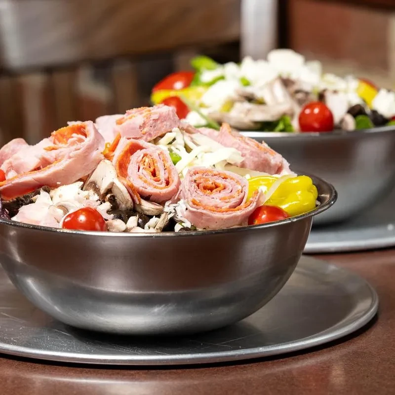 Two large salads with deli meat, cheese, and vegetables in steel bowls.