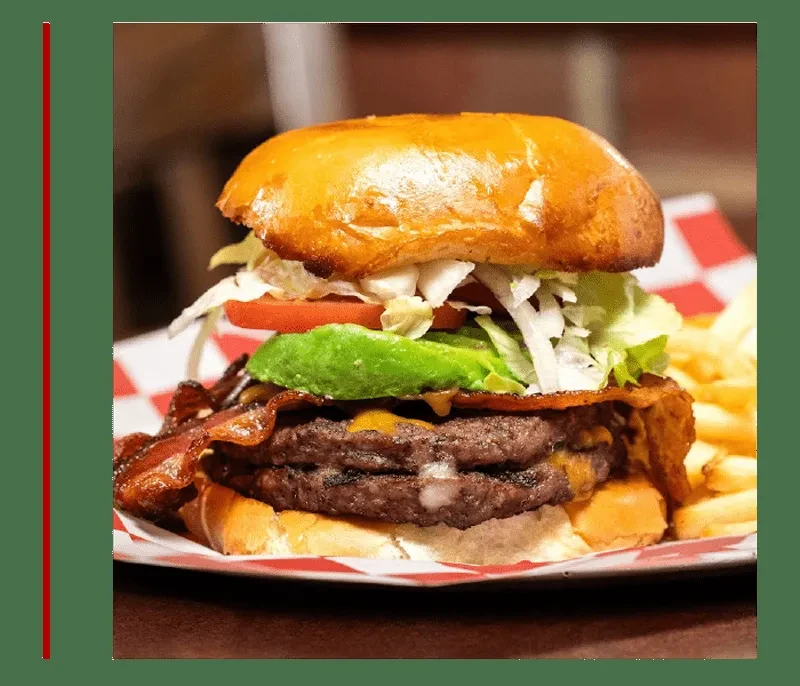 Double-patty burger with bacon, avocado, fries, on checkered diner paper.