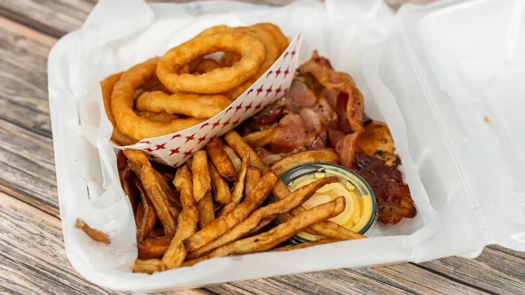 Take-out container with fried onion rings, french fries, meat, and dipping sauce.