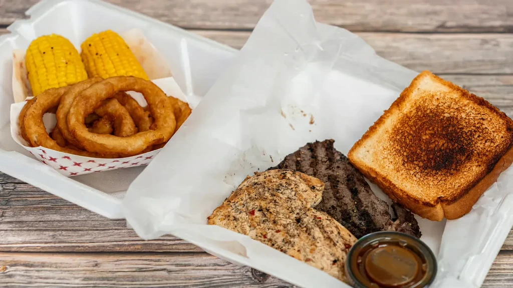 Two takeout containers hold grilled chicken, meat, corn, onion rings, and toast.