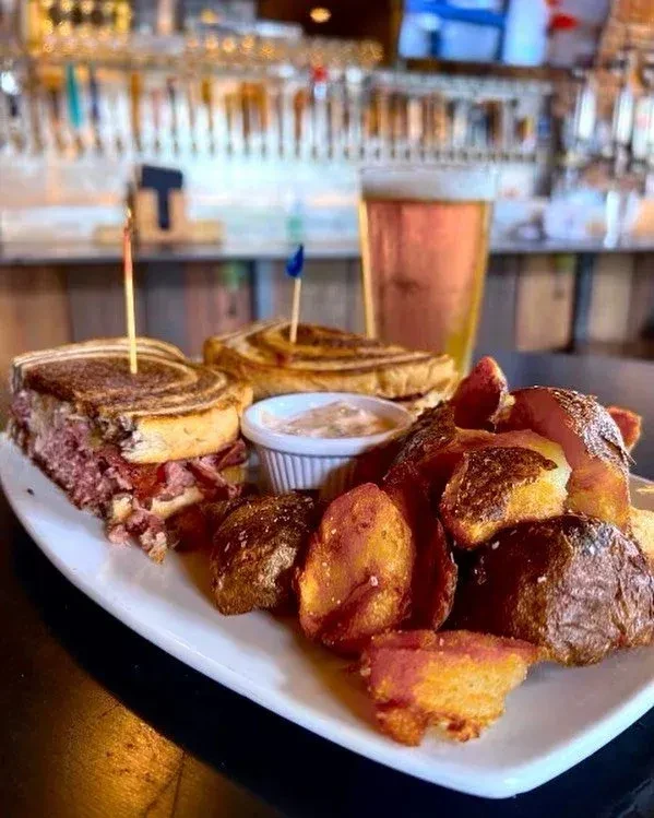 Grilled Reuben sandwich, potato wedges, creamy dip, and beer at a bar.