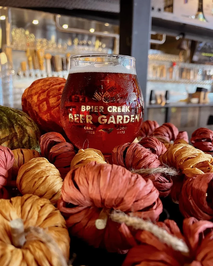 Condensation-covered amber beer glass surrounded by fall pumpkin decorations.