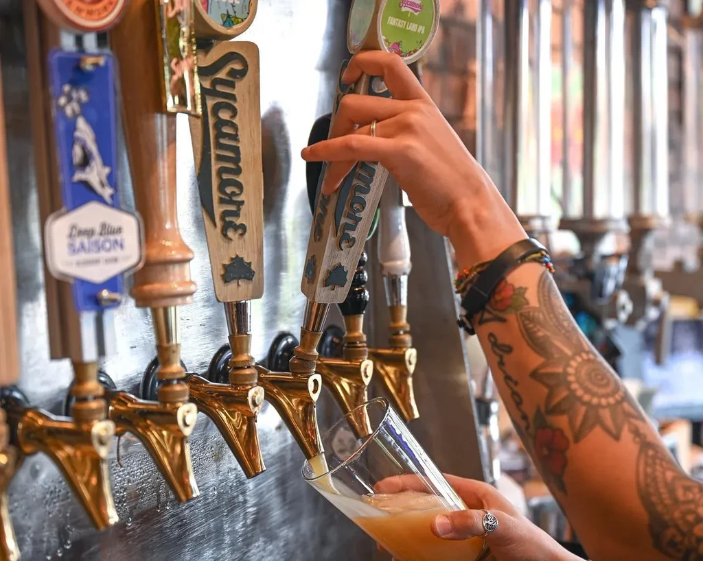 Tattooed arm pouring frothy beer from one of several bar taps.