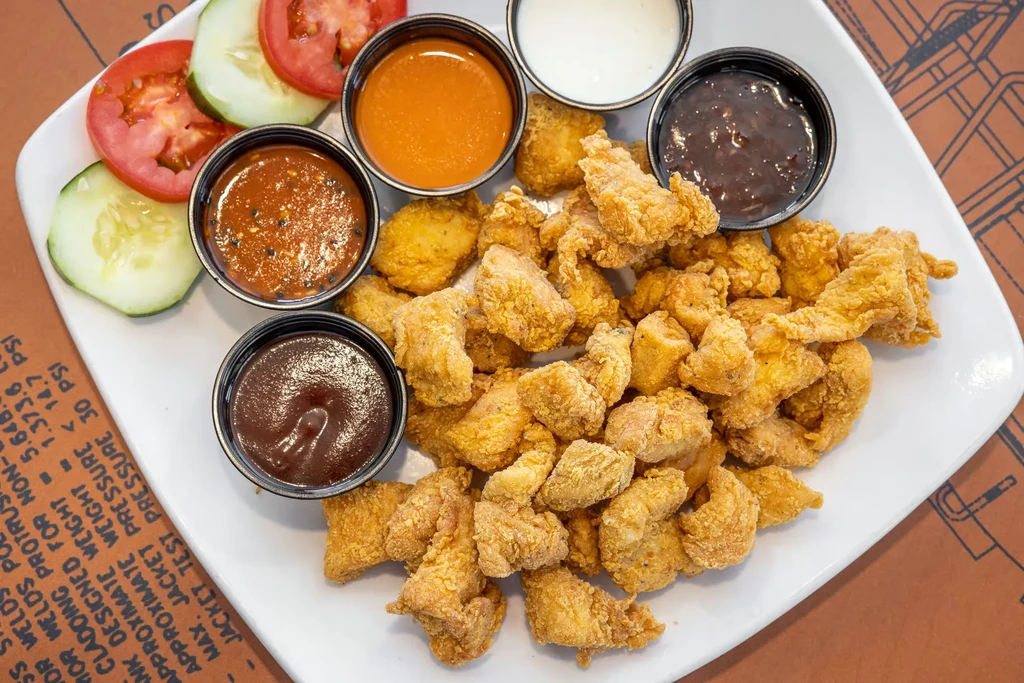 Bite-sized fried chicken, five sauces, cucumber, and tomato on a plate.