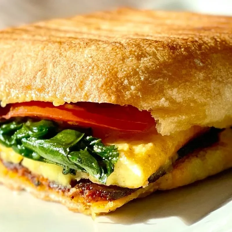 Golden-brown toasted sandwich with tomato, greens, and creamy yellow layer.