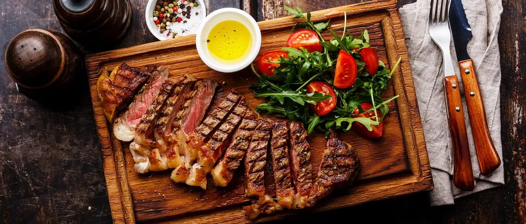 Sliced medium-rare steak, arugula salad, and spices on rustic wood.