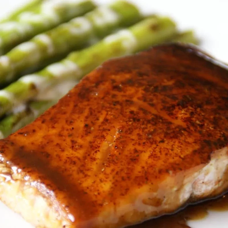 Close-up of seasoned salmon with dark, glossy sauce and blurred asparagus.
