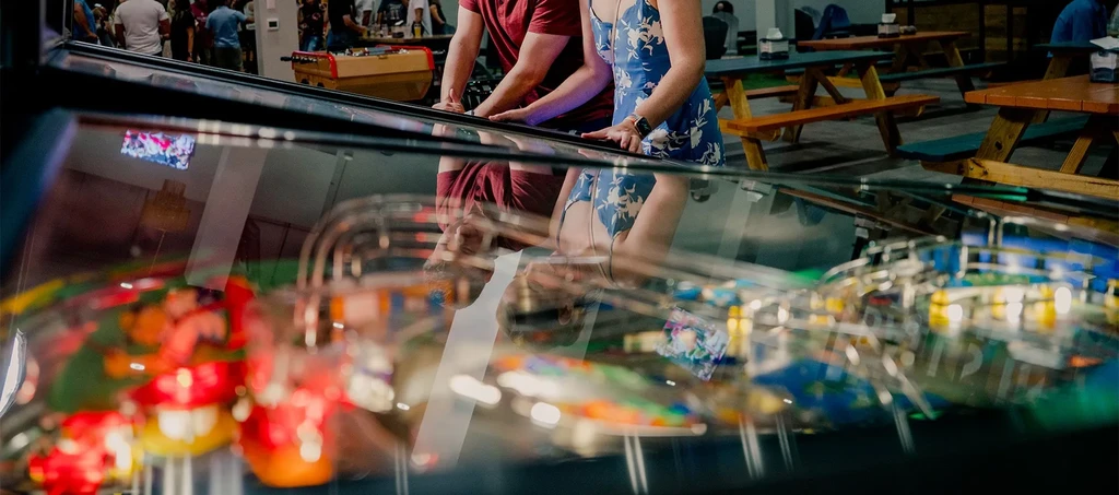 Two people play a pinball machine in a lively arcade bar.
