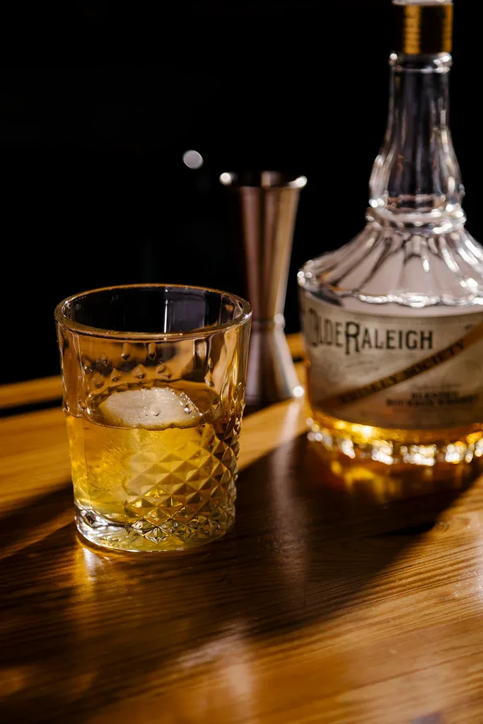 Patterned glass of whiskey with ice, bottle, jigger on wood.