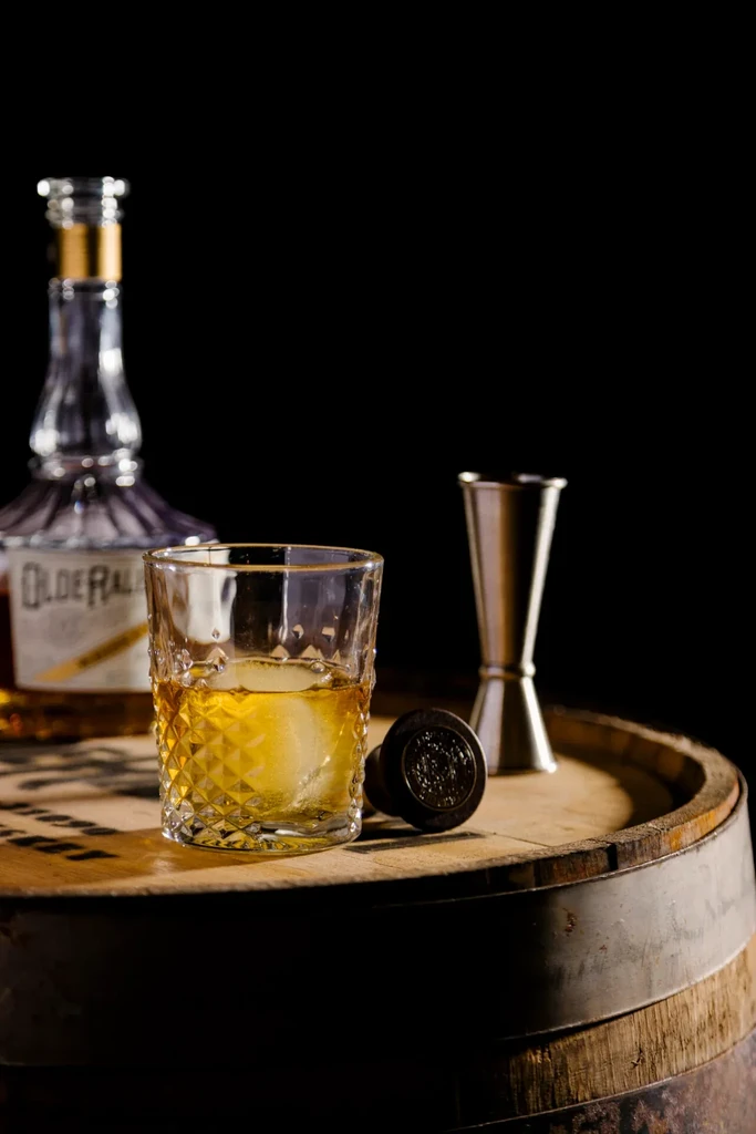 Whiskey glass with ice, bottle, jigger, stopper on rustic wooden barrel.