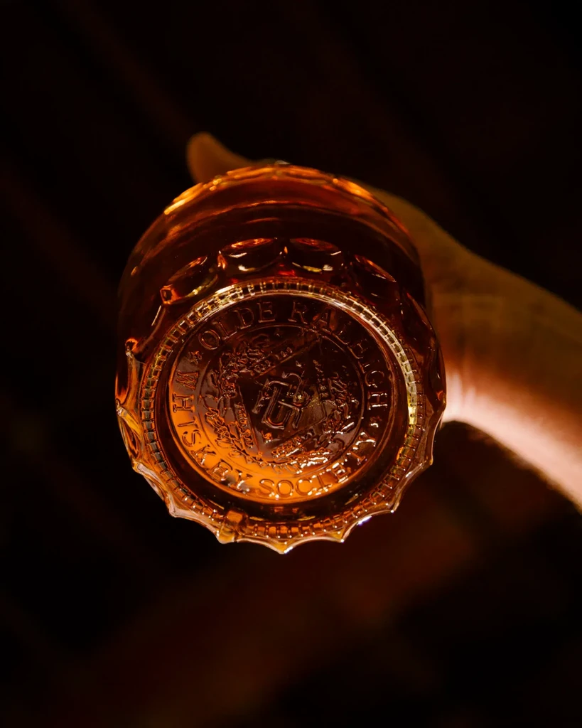 Hand holding amber whiskey glass showing 'OLDE RALEIGH WHISKEY SOCIETY' seal.