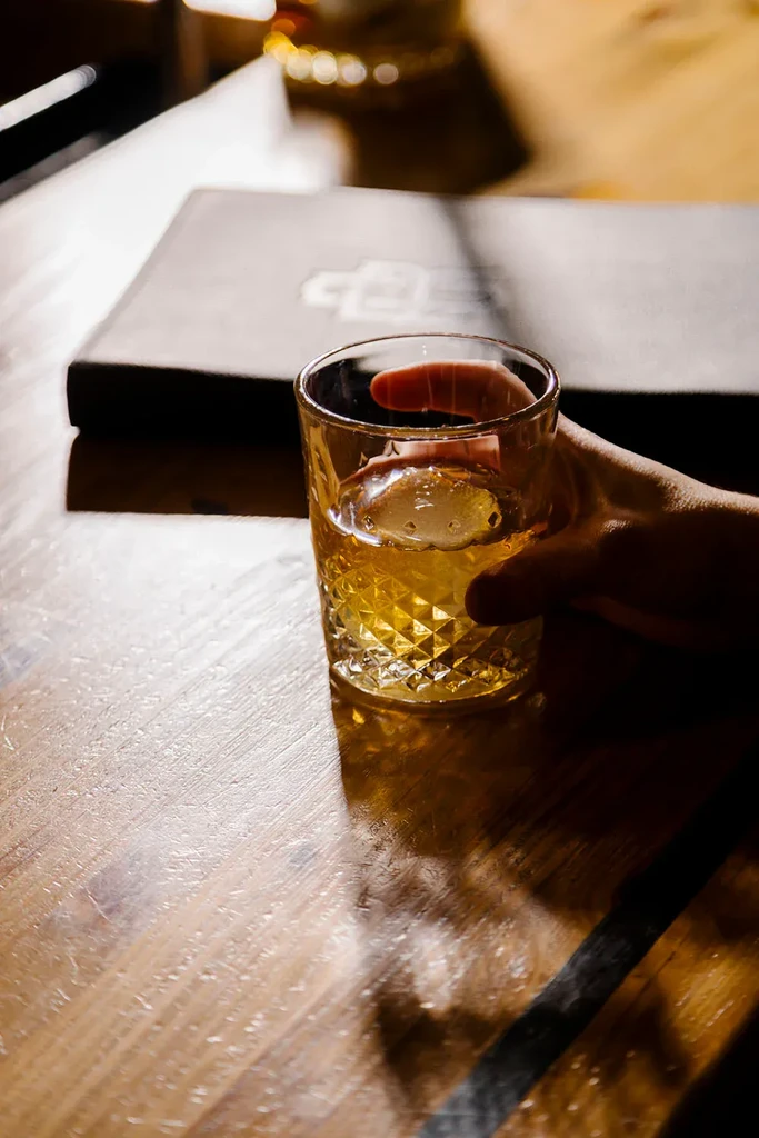 Hand holding glass of whiskey with spherical ice on wooden table.