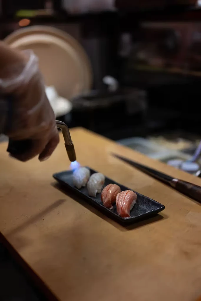 Gloved hand torches four nigiri sushi pieces on a black plate.
