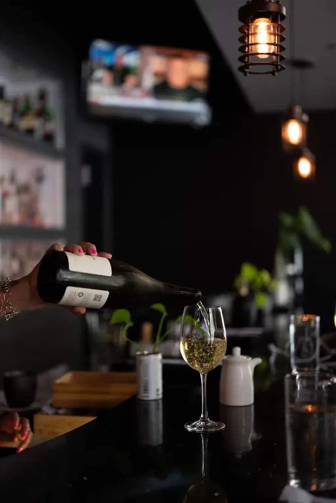 Hand with pink nails pours white wine into glass at dark bar.