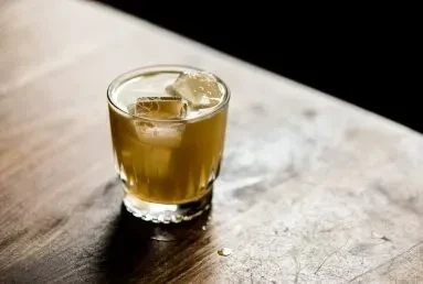 Amber drink with ice in a faceted tumbler on a worn table.