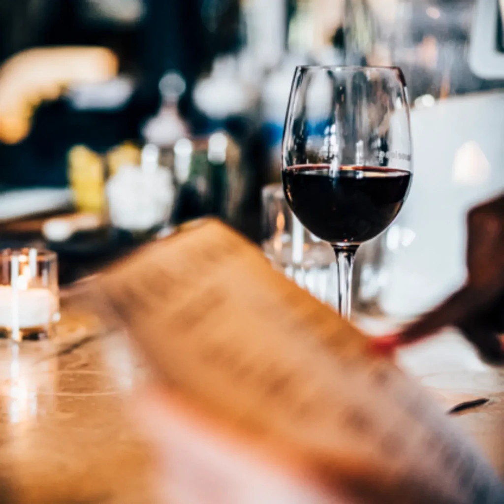 Red wine glass, blurred menu, lit candle, and faint hand.