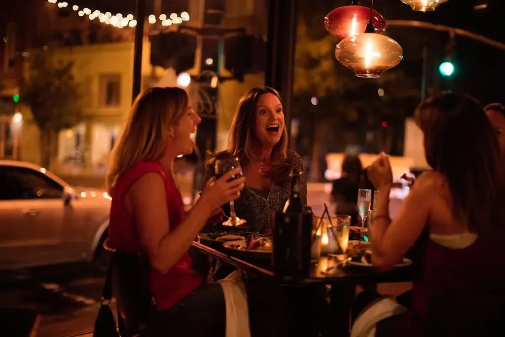 Three women laughing and dining at a lively restaurant at night.