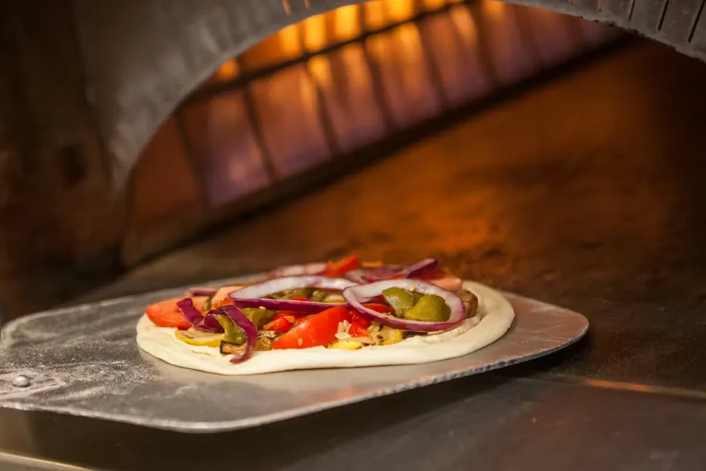 Uncooked vegetable pizza on peel entering a flame-lit oven.