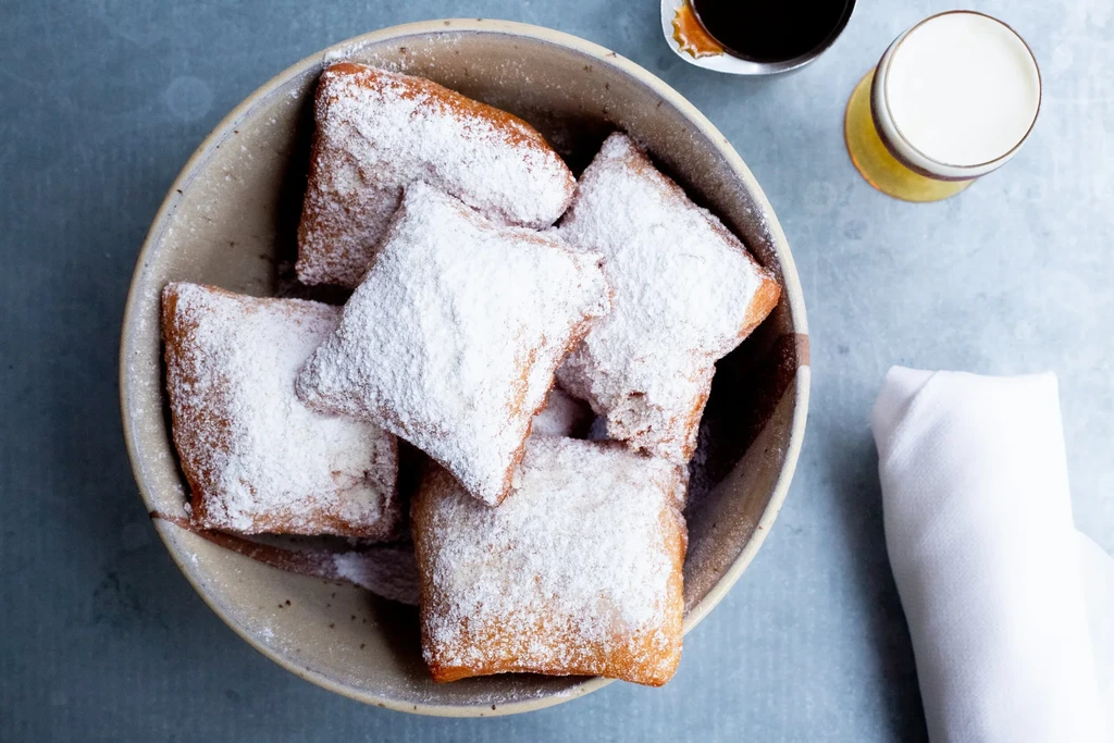 Bowl of powdered sugar beignets next to coffee and a foamy beverage.