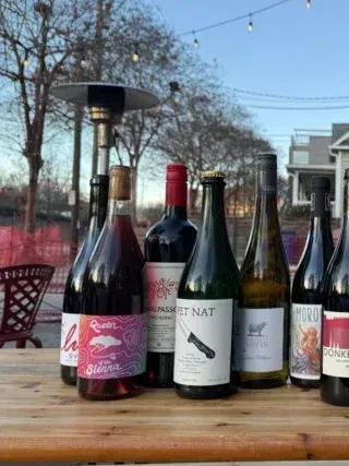 Eight diverse wine and liquor bottles on a wooden table outdoors.